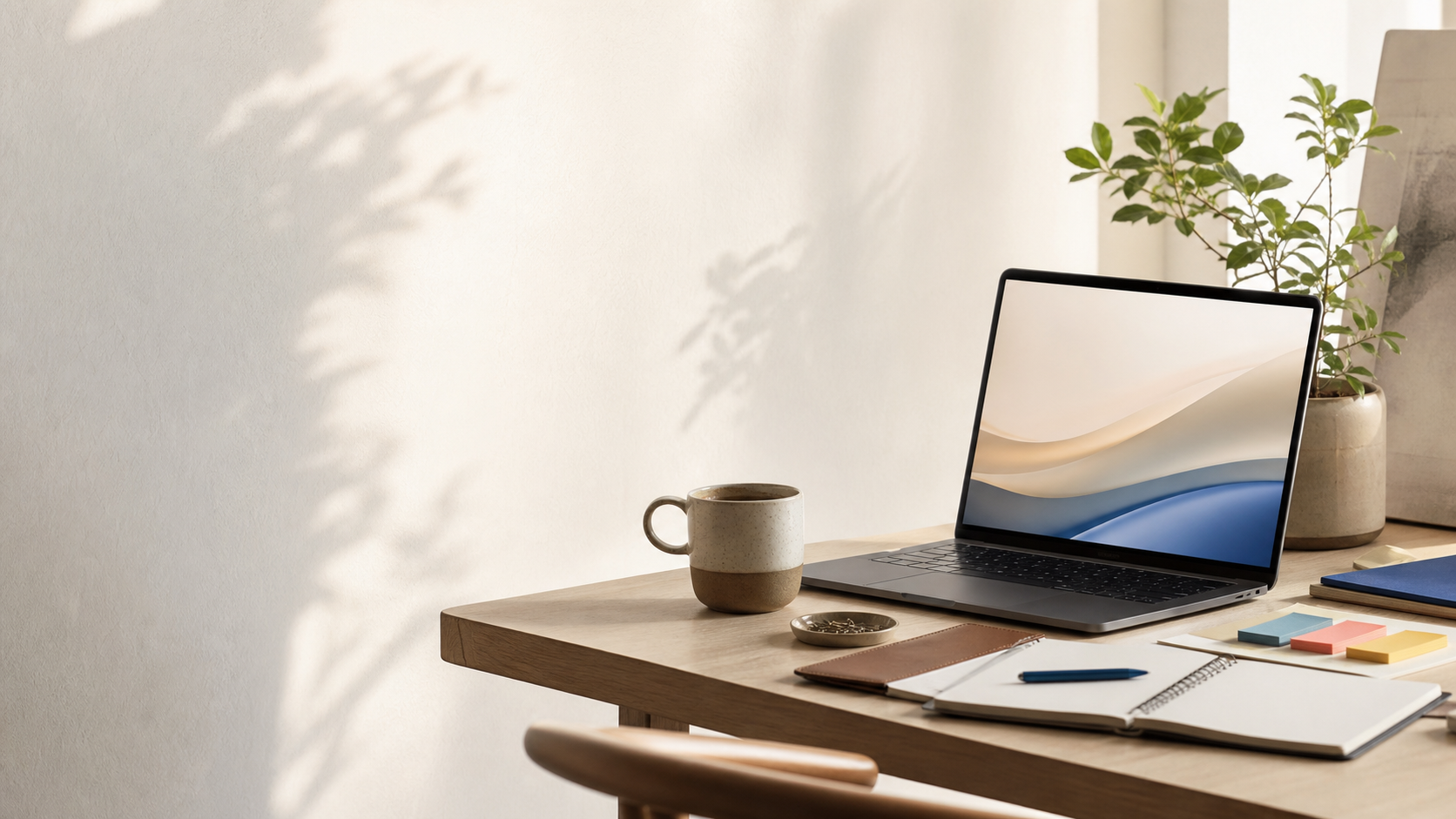 A warm desk with a laptop, notebook, coffee, and plant in soft daylight.